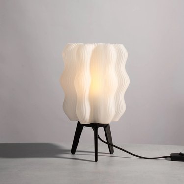 A wavy, 3D-printed table lamp with a white translucent shade and three-legged black base on a white background.