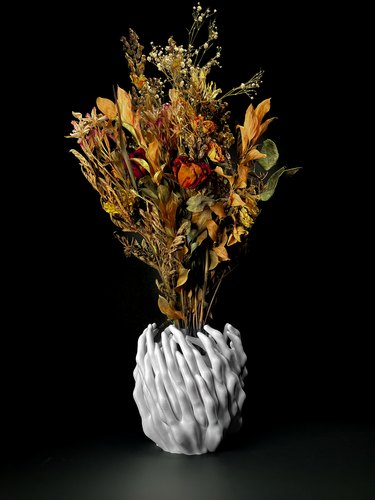 A white base holds an autumnal bouquet of roses and other flowers. The surface of the vase is jagged, and resembles fingers that have been woven together.