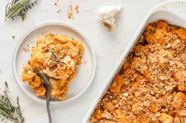 A casserole dish filled with the squash bake next to a plate with a piece of the bake on it and a head of garlic on a white marble counter.