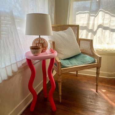 The image shows a corner of a brightly lit room.  A side table is arranged next to a wicker chair. The side table has three squiggly hot pink legs and a circular bubblegum pink surface. A desert-colored ceramic mug and  a round terracotta lamp are placed on the side table.