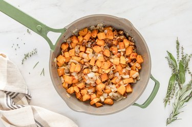 The onion, butternut squash, mushrooms, cinnamon, and minced garlic in a green frying pan on a white marble countertop.