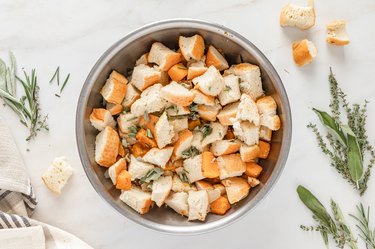 Tossed cubed bread, butternut squash mixture, and chopped sage, rosemary, and thyme in a metal bowl on a white marble countertop.