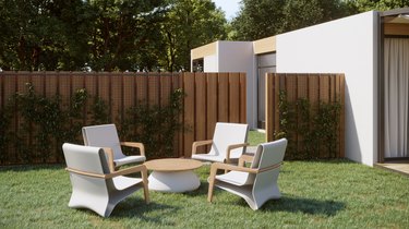 Four chairs are arranged about a rounded coffee table. The chairs are made of white plastic and wood, with a wavy design. The coffee table has a white plastic base and a circular wooden surface. The furniture is arranged outside, in a grassy backyard. There is a fence and trees in the background.