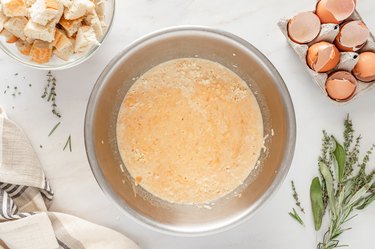 Half-and-half, broth, pumpkin puree, salt, black pepper, smoked gouda cheese, and eggs whisked together in a metal bowl on a white marble countertop.