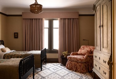 Bedroom with two twin beds, muted yellow wallpaper, floral side chair and mauve curtains.