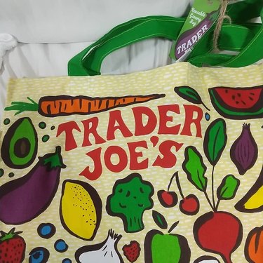 Zoomed in picture of a Trader Joe's reusable tote with the logo and vegetable drawings like eggplant, avocado, broccoli, and cherries.