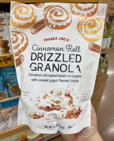 A white bag of Trader Joe's cinnamon roll drizzled granola. There are drawings on cinnamon rolls and cinnamon sticks at the top, with an image of the granola itself at the bottom. It's a granola cluster with yogurt drizzle on top.