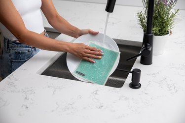 A woman wearing a white top and jeans washes a white plate with a teal swish cloth in a kitchen sink. The kitchen counter is white marble, and the faucet is black. There is a lavender plant resting next to the faucet.