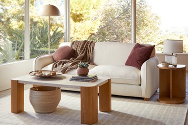 A living room with large glass windows showcasing various plants. There is a white sofa topped with a dark red pillow and brown throw, along with a wood and travertine coffee table topped with a plant and decorative tray.