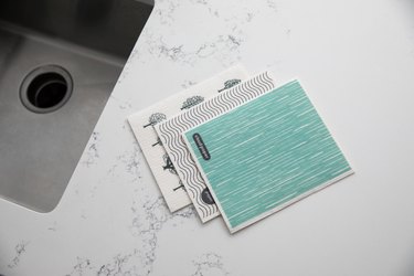 Three swish cloths rest in a pile on a white marble kitchen counter next to a sink. The cloth on top is teal with thin white lines throughout, the second cloth is white with horizontal, wavy lines and the third cloth is white with images of trees on it.