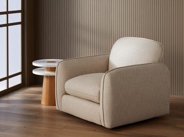 A pillowy armchair in a flax beige fabric next to a side table with a wood base and two layers of marble for tabletops.