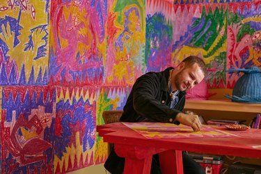 Daniel Valero drawing at a red table in front of a colorful background of purples, yellows, reds, blues, and greens.