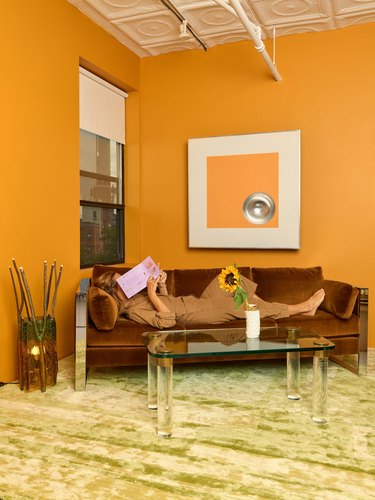An orange wall with a woman laying on a brown couch holding a book in front of her face. There is a glass coffee table with a sunflower in a vase on it.