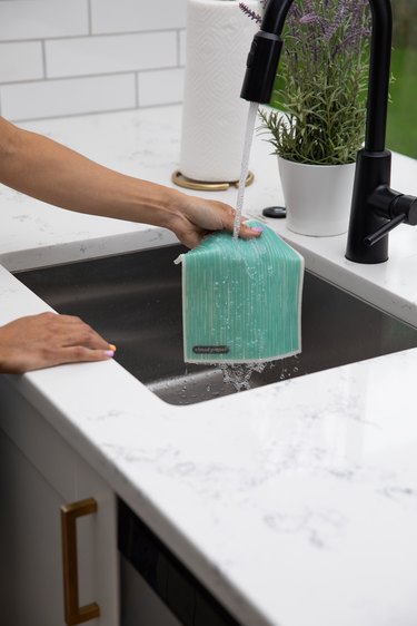 A woman's hand holding a teal swish cloth underneath running water in a kitchen sink. The faucet is black and the kitchen counter is white marble, with a lavender plant close by, as well as a roll of paper towels.