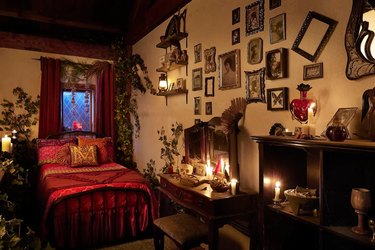 Mary Sanderson's loft in the Hocus Pocus Cottage with a red bedding, a wooden desk with candles, and crooked picture frames covering the wall.