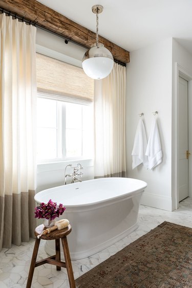 White bathroom with soak tub, large pendant light, oriental rug, curtains.