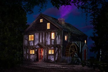 Exterior of the Hocus Pocus Cottage at night with glowing yellow windows and purple smoke coming out of the chimney.
