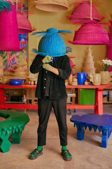 A man dressed in black holding a bright blue wicker basket over his head in front of a studio composed of a work station, other wicker baskets, and colorful benches.