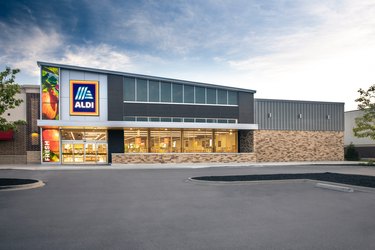 The exterior of an Aldi store. The building has a slanted roof, with the Aldi logo at the top on the right side of the building, with an image of apples on a tree that cover the whole right side of the building, with the word "fresh" on the bottom.