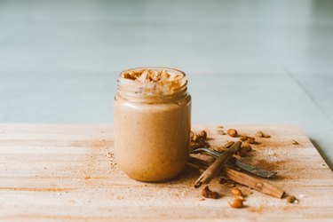 A jar of homemade peanut butter on a wood cutting board with cinnamon sticks and peanuts.