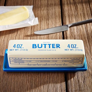 A stick of butter being stored on a blue butter dish. There is a silver butter knife in the background on the wood tabletop.