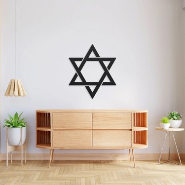 A black, minimalist Star of David hanging on a white wall over a wooden console on wooden floors.