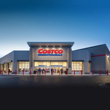 The exterior of a Costco at sunset. The store front has a large sign that says "Costco Wholesale" and is lit from above. A few people can be seen in front of the store.