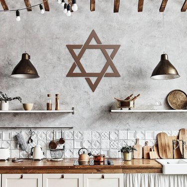 Metal Star of David hanging on a kitchen wall next to bronze lights, white shelves, and a wooden countertop.
