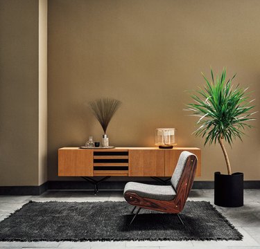 A walnut credenza behind an armless upholstered gray chair on a black fringe rug next to a leafy small palm tree.