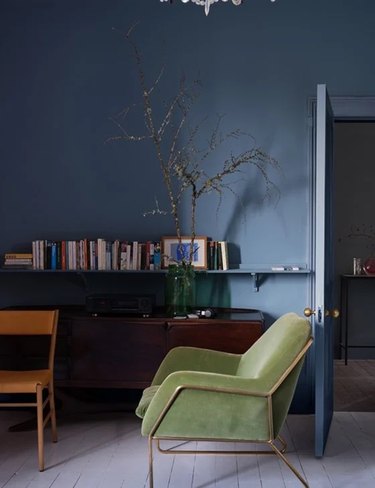 Farrow & Ball's medium-light blue paint color on an office wall behind a green armchair and wall shelf covered in books.