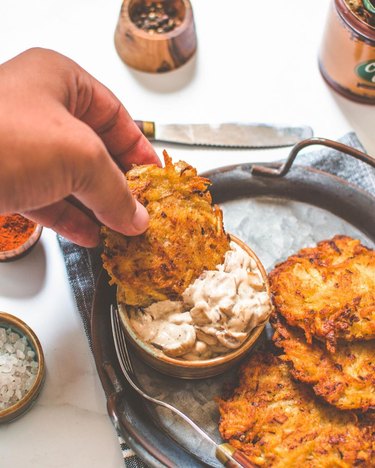 Hand dipping vegan latkes in a caramelized onion dip on a silver tray.