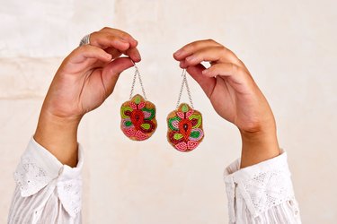Two hands holding up a pair of beaded earrings in front of a beige wall.