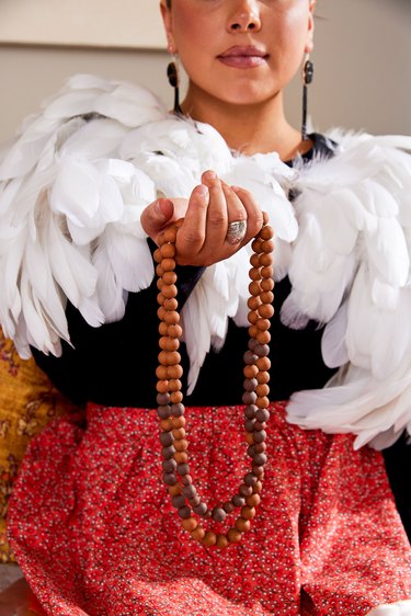 A woman in a black shirt, red skirt, and white feather boa holding out a brown beaded necklace towards the camera.