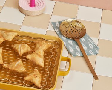 Samosas on top of a gold cooling rack in a turmeric-colored oven pan next to a slotted spoon on a blue and white napkin.