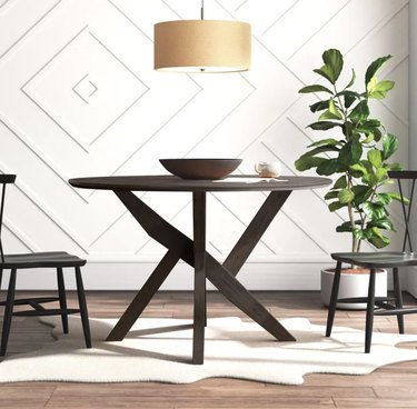 Image of a brown round dining table and two chairs. There's a bowl in the center of the table and a plant in the corner of the room.