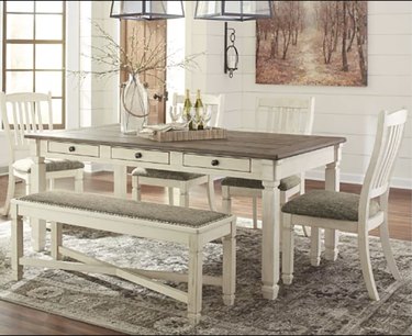 Image of distressed white farmhouse dining table set with dark wood tabletop. The table is surrounded by four matcching chairs and a bench.