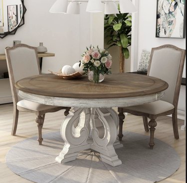 Image of a distressed round dining table with a dark wood tabletop. There are two upholstered chairs around the table. A small vase filled with roses and a tray with round while balls sit on the table.