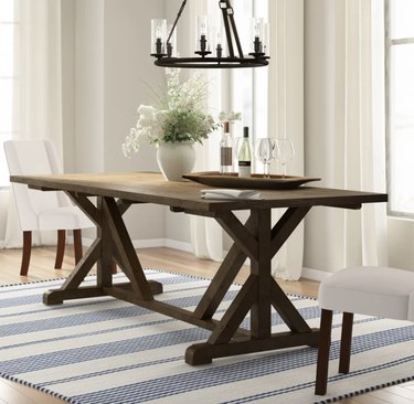 Image of a large farmhouse table with a trestles. The table is a dark wood color and accessorized with vases and wine and wine glasses.