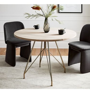 Image of a mid-century modern round dining table with brass colored legs. Two black chairs surround the table, and a vase with flowers and two cups sit on the table.