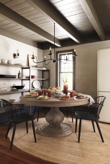 Image of a round farmhouse dining table with a large pedestal. The table is in a kitcchen, situated on a burlap rug, surrounded by black chairs and it's set for a meal.