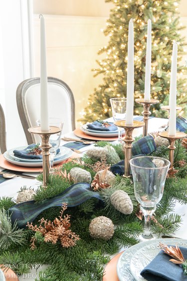 Christmas table spread with garland, blue and greed plaid ribbon, and copper candlesticks and pinecones.