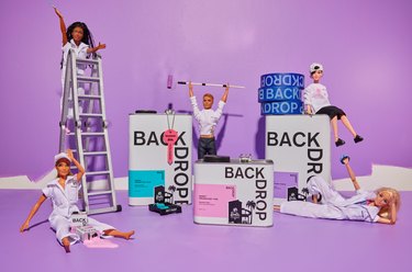 Barbie Dolls over Backdrop paint bottles with a ladder and paintbrushes on a purple background.