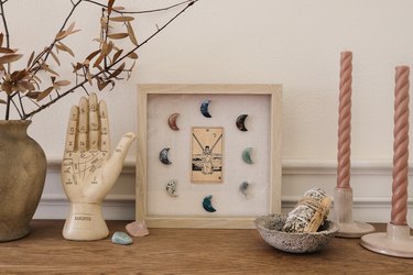 Shadow box with tarot card and moon-shaped crystals on table with palmistry hand, sage, and candles