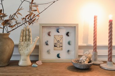 Shadow box with tarot card and moon-shaped crystals on table with palmistry hand, sage, and candles