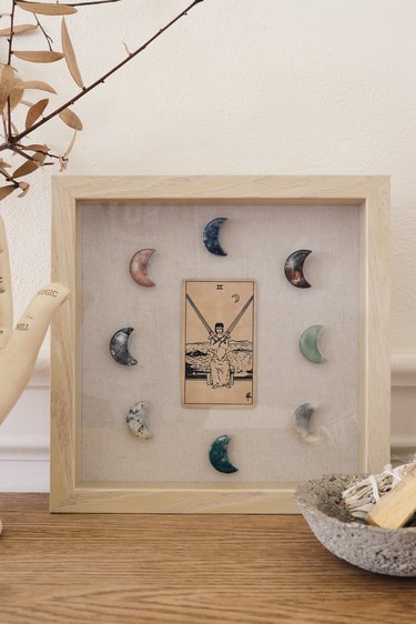 Shadow box with tarot card and moon-shaped crystals on table with palmistry hand, sage, and candles