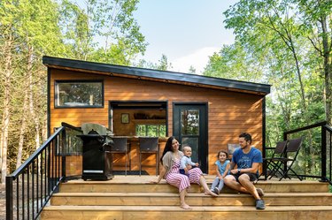 Outdoor shot of modern rustic wood-paneled house with outdoor deck with parents and children against trees and blue sky