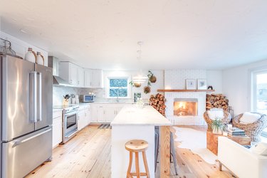 a large kitchen with an island, white cabinets, and a rustic pine floor flows into a living-room area with a fireplace