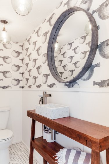 a bathroom with a basin sink on top of a crafted wood vanity, a round mirror, and wallpaper with a puffer fish pattern