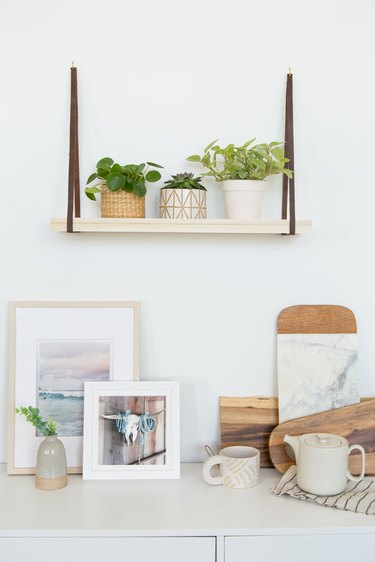 Hanging plant shelf with plants in planters, and made from wood and leather over artworks, mugs, and wood cutting boards
