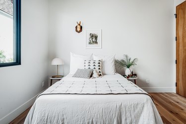 White bedding with patterned pillows and a stuffed animal. A white half dome table lamp. A geometric vase with palm leaves, and wood floor.